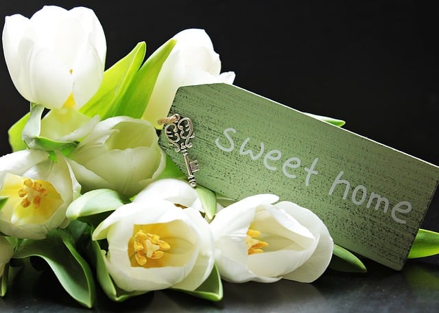 A bouquet of white tulips lies next to a vintage-style key and a green wooden sign that says "sweet home" on a dark surface, capturing the excitement of moving house and making the experience stress-free.