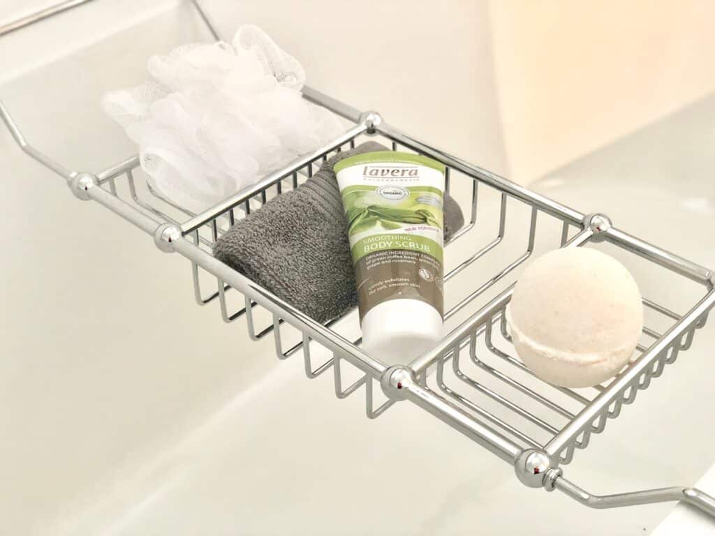A metal bath caddy holds a white bath puff, a dark grey flannel, a tube of Lavera body scrub, and a round white bath bomb—essentials for nightly rituals and stress relief over a relaxing soak in the bath.