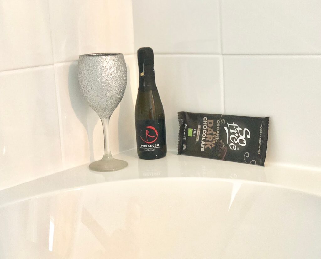 A glittery silver wine glass, a small bottle of prosecco, and a bar of So Free dark chocolate are arranged on the edge of a white bath—perfect companions for nightly rituals and stress relief.