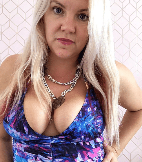 A woman with long blonde hair wearing a blue and purple patterned top, reminiscent of stylish swimming costumes for women, and a chunky silver chain necklace stands in front of a geometric-patterned pale pink wall, looking directly at the camera.