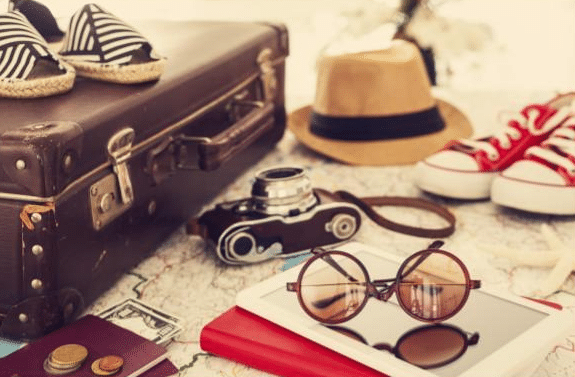 A vintage suitcase, striped shoes, a map, a camera, a hat, red trainers, sunglasses, a tablet, coins, and a passport are arranged as the perfect holiday packing list for your next adventure.