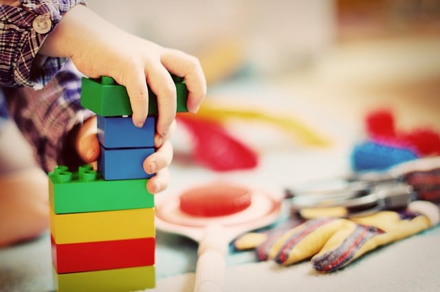 A childminder watches as a child’s hand stacks colourful plastic building blocks. Various toys are scattered in the blurred background, creating a playful and creative scene.