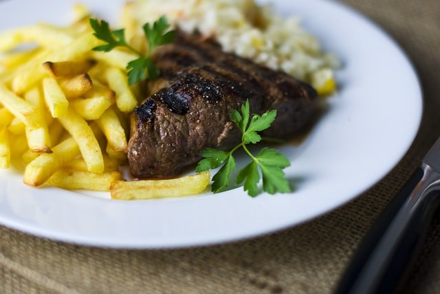 A white plate with grilled steak pieces, golden chips in classic bistro steak frites style, creamy rice, and a sprig of parsley, set on a hessian surface beside a knife.