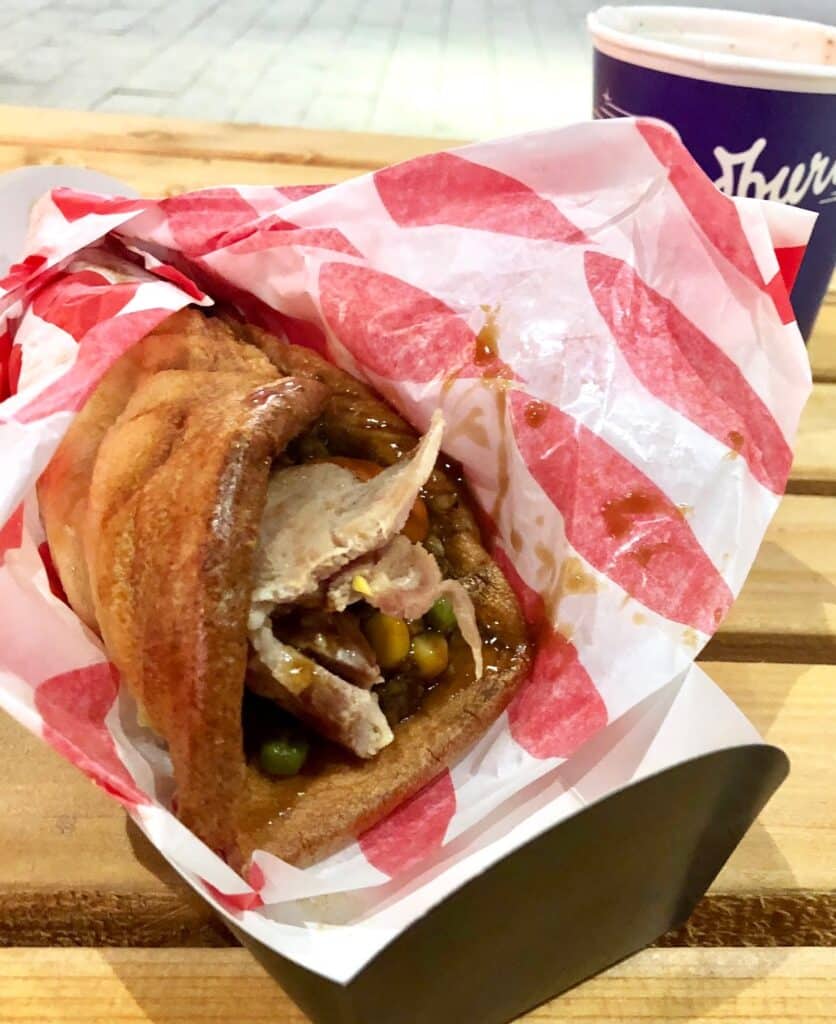 A close-up of a Yorkshire pudding wrap filled with roast meat, peas, and gravy, wrapped in red and white paper on a wooden table at the Yorkshire Festival, with a drinks cup in the background.