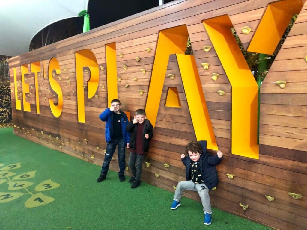 Three young boys pose and smile in front of a large wooden wall with bright yellow 3D letters spelling "LET'S PLAY" at the Yorkshire Festival. The wall has climbing holds, and the ground is covered in green safety flooring.