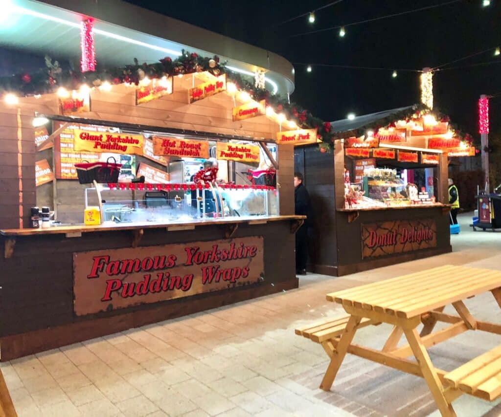 A festive festival market at night with wooden stalls selling Yorkshire pudding wraps and doughnuts. The stalls are decorated with lights and garlands, whilst a wooden picnic table sits in the foreground, capturing the spirit of White Rose Yorkshire.