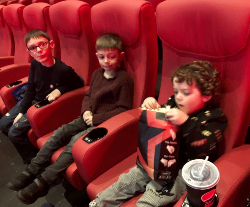 Three young children sit in red cinema seats at the White Rose Yorkshire Festival. The child on the right enjoys popcorn from a large bag, whilst the others watch. A drinks cup with a straw rests in the cup holder.