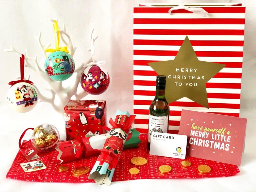 A festive arrangement inspired by the White Rose Yorkshire Festival, featuring a red and white striped gift bag, Christmas baubles on a white tree, wrapped presents, a small wine bottle, gold chocolate coins, a gift card, crackers, and a Christmas card.