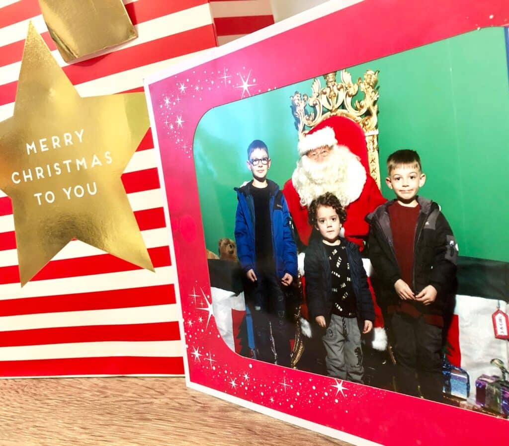 A framed photo of three young boys posing with Father Christmas is placed on a wooden surface next to a large red and white striped gift bag, adding charm to your Festive Festival or White Rose Yorkshire celebration.