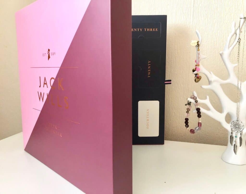 A pink and purple Jack Wills gift box stands on a table next to a black Advent Calendar, adding festive flair to your holiday countdown beside a white jewellery tree holding bracelets and earrings.