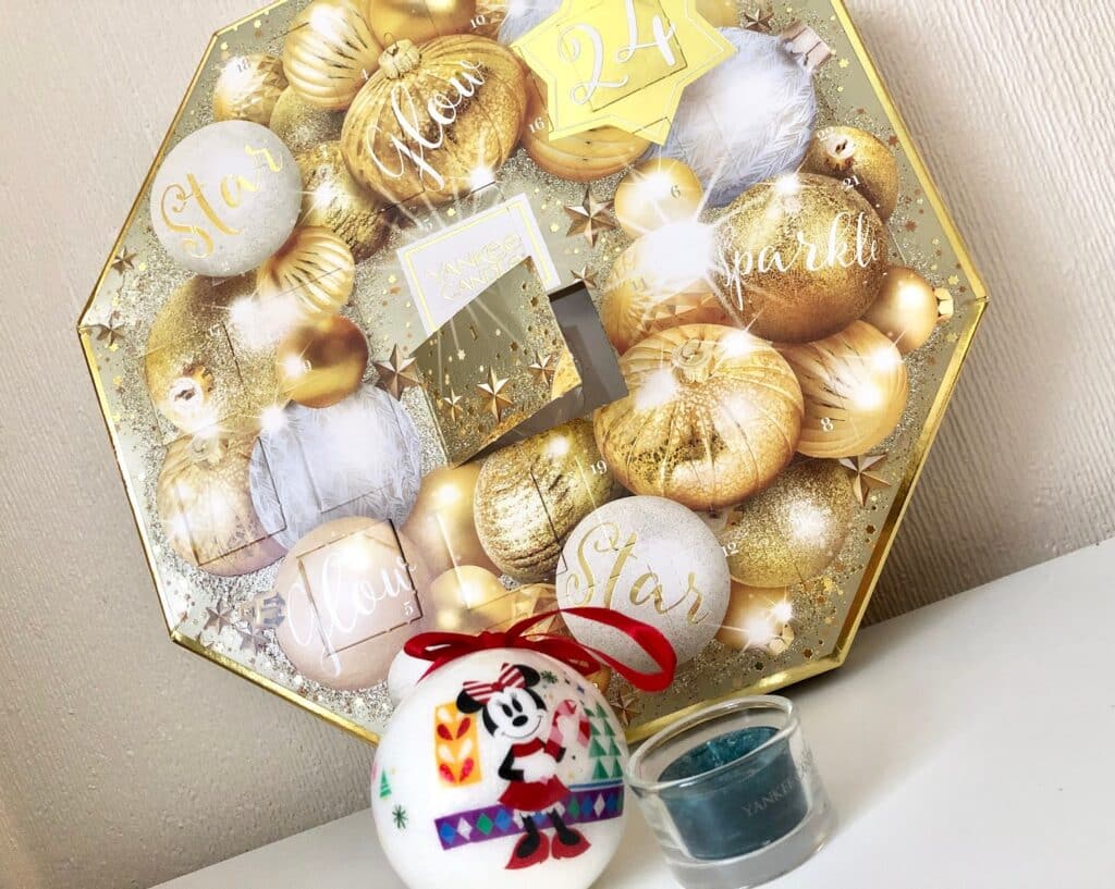 A gold and white Advent Calendar decorated with baubles stands upright, bringing festive Holidays cheer. In front, a round white ornament with a cartoon mouse and red ribbon sits by a small blue Yankee Candle in a glass holder.
