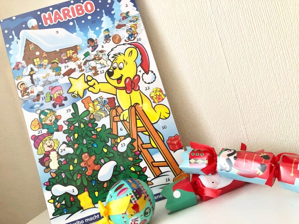A Haribo advent calendar featuring a cartoon bear in a Santa hat decorating a Christmas tree, perfect for family holidays. Colourful holiday crackers and a decorated bauble add festivity to the scene.