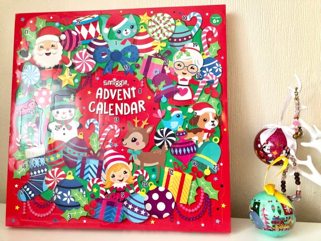 A colourful Smiggle Advent calendar with festive cartoon characters, gifts, and decorations on a red background sits next to a white reindeer ornament holding two decorated Christmas baubles—a perfect touch for holiday family fun.