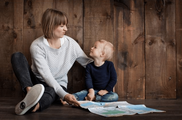 A woman and a young child sit on the floor against a wooden wall, smiling at each other as they look at a map—sharing a comforting moment that reminds us how to help children incontinence with warmth and support.