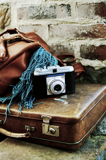 A vintage camera rests atop an old brown suitcase, next to a leather bag and a blue fringed scarf—an inspiring scene for first-time cruisers seeking a stylish cruise packing guide against a rustic brick wall.