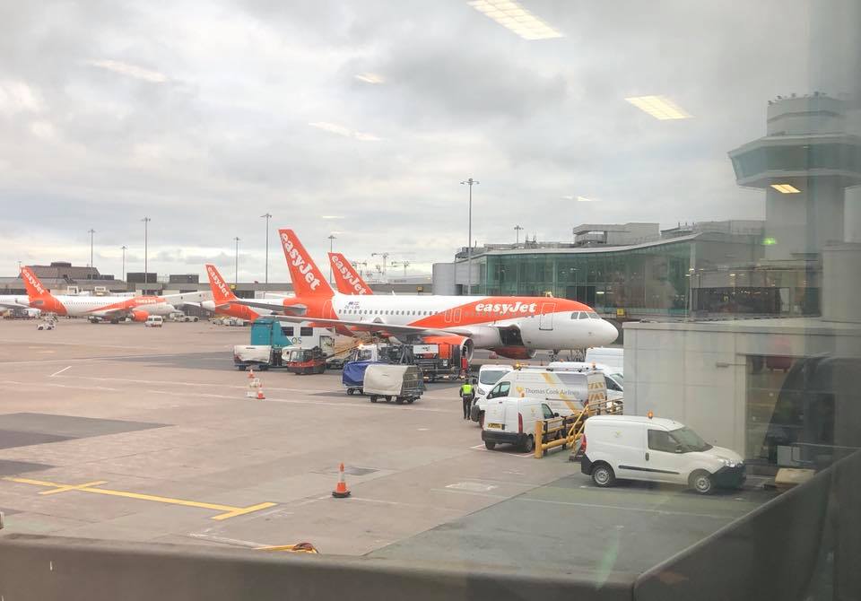 A view through a window at an airport shows several easyJet aeroplanes parked at gates, with ground vehicles and staff working around them under a cloudy sky—capturing the excitement of going abroad.