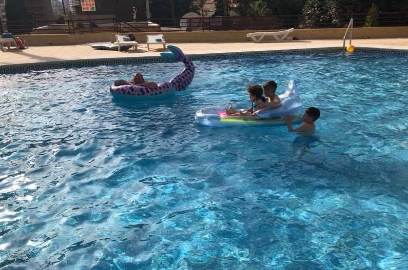 Three children sit on an inflatable raft in a pool, whilst another child relaxes on a mermaid tail float nearby. The scene feels like going abroad, with clear blue water and sunlight sparkling on the surface.