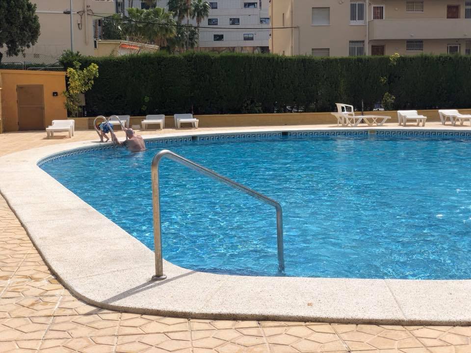 A person sits alone at the edge of an outdoor swimming pool, dipping their feet in the water, lost in thought, perhaps dreaming of going abroad. Empty sun loungers surround the pool, with tall hedges and blocks of flats in the background.