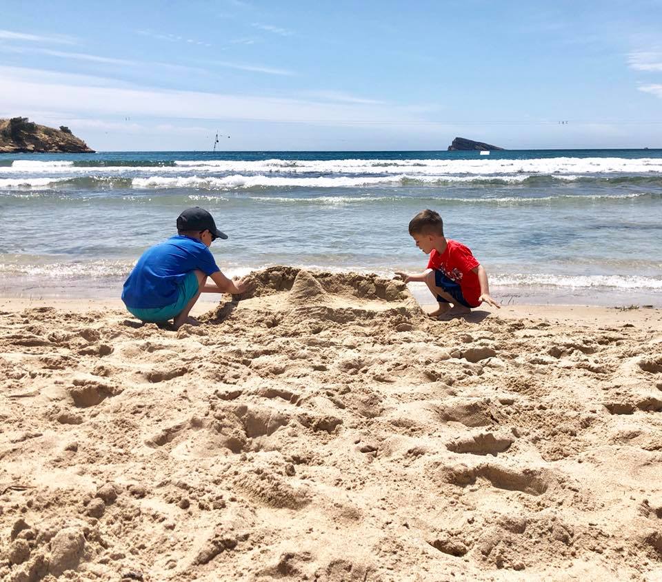Two young boys play in the sand near the shoreline, building a sandcastle together on a sunny beach, dreaming of going abroad whilst waves lap and a small island appears in the background.