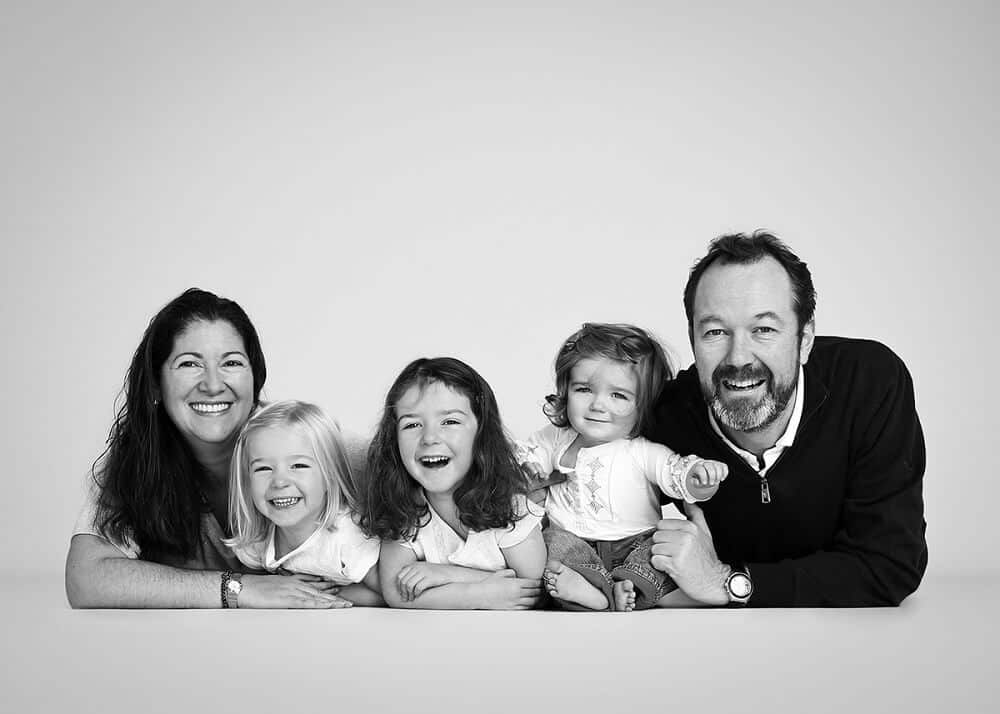 A black and white photo of a smiling family of five, with two adults and three young children, lying on the floor together against a plain background—a perfect family photography experience.