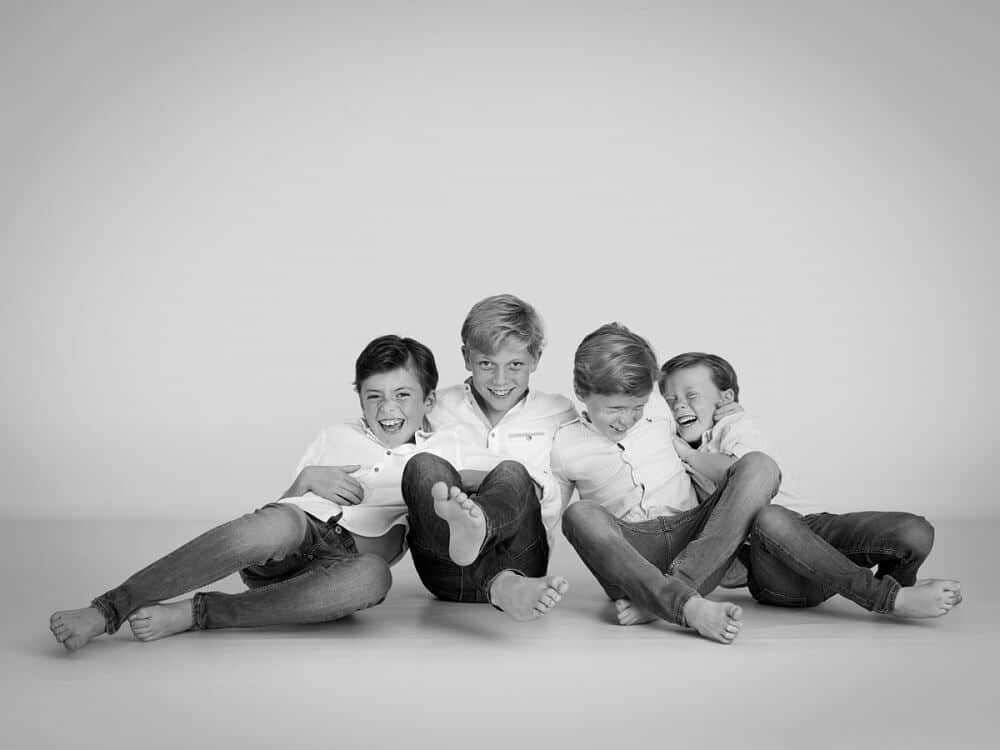Four young boys sit barefoot on the floor, wearing matching white shirts and jeans. Laughing and playful, they lean into each other in this black and white family photography session—capturing a joyful moment perfect for a Christmas present.