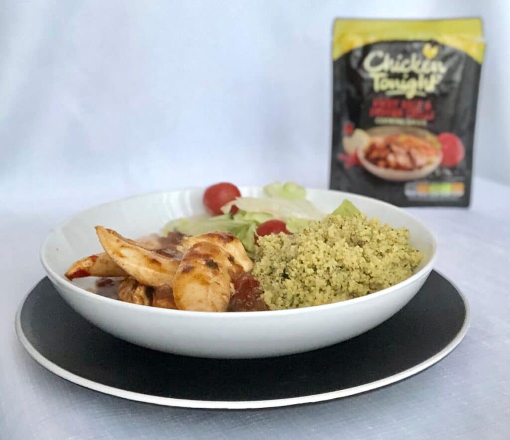 A white bowl holds cooked chicken in sauce, green couscous, and salad with cherry tomatoes. In the background, a "Chicken Tonight" packet promises to banish midweek monotony. The bowl sits on a black plate.