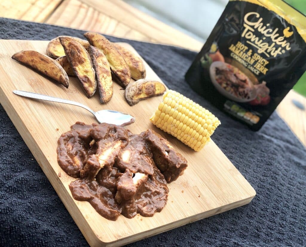 A wooden board with potato wedges, a corn cob, and chicken in a dark sauce beside a spoon—perfect for Midweek Meals. A yellow and black "Chicken Tonight" sauce packet is in the background on a black cloth to help rid monotony from your tea-time routine.