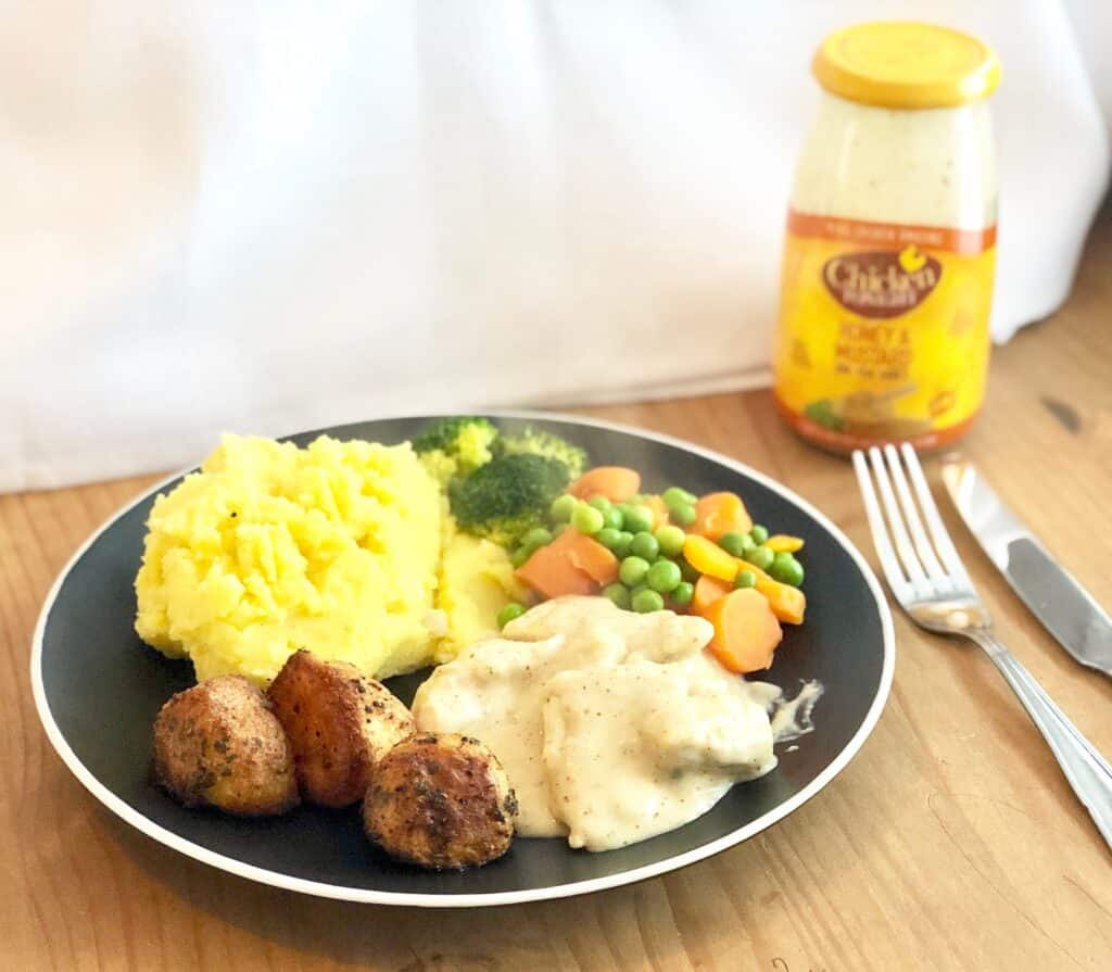 A plate of mashed potatoes, broccoli, mixed peas and carrots, browned meatballs, and chicken in white sauce is perfect for a cosy midweek dinner. A jar of chicken stock sits in the background beside a fork and knife.