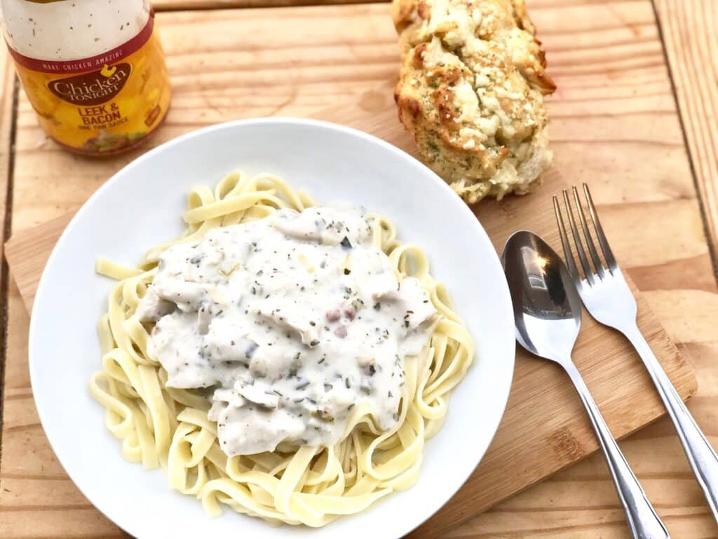 A plate of fettuccine with creamy white sauce and herbs, a bread roll, cutlery on a wooden board, and a jar of leek and bacon simmer sauce—perfect inspiration for easy chicken recipes or your next midweek dinner.