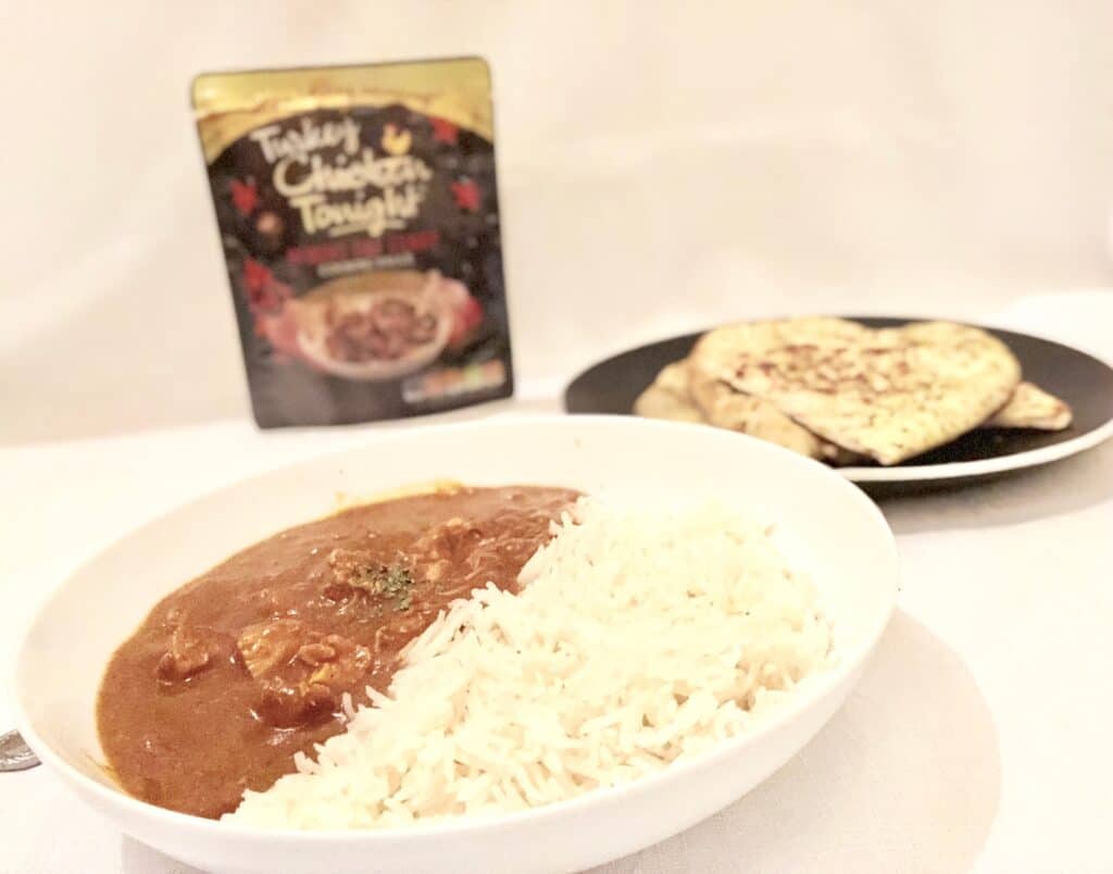 A white bowl with chicken curry and rice sits in the foreground, making the perfect midweek dinner, with a plate of naan bread and a packaged Chicken Tonight curry sauce in the background on a white surface.