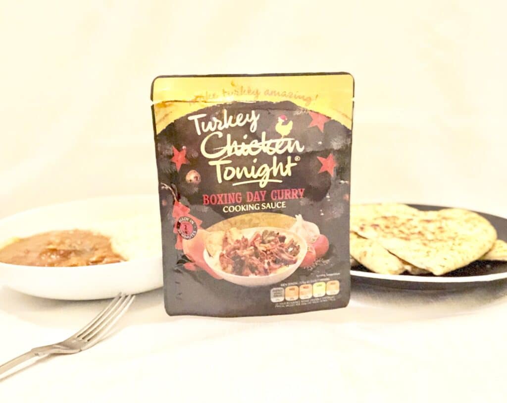 A packet of Chicken Tonight "Turkey Chicken Tonight Boxing Day Curry Cooking Sauce" stands between a bowl of curry with a fork and a plate of naan bread, ready to break the Midweek Monotony on a white surface.