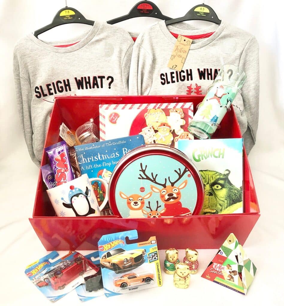 A festive Christmas Eve Box contains matching "Sleigh What?" shirts, a reindeer tin, Christmas books, Hot Wheels cars, snacks, a tumbler, stickers, and other holiday-themed treats—all arranged against a white background.