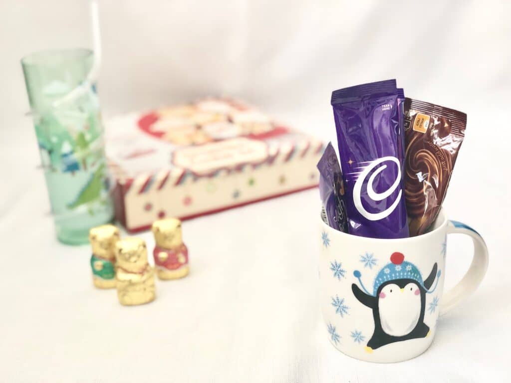 A festive mug with a penguin design holds hot chocolate sachets. Nearby are three gold-wrapped chocolate bears, a Christmas Eve Box, and a light blue glass with a straw, all set against a white background.