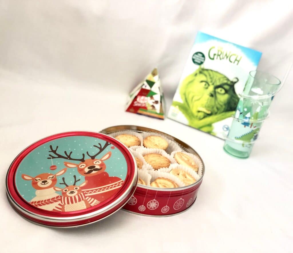 A festive tin with reindeer design holds butter biscuits, perfect for a Christmas Eve Box. Nearby are a Grinch DVD, a geometric holiday gift box, and a tall glass with a snowman, all set on a white background.