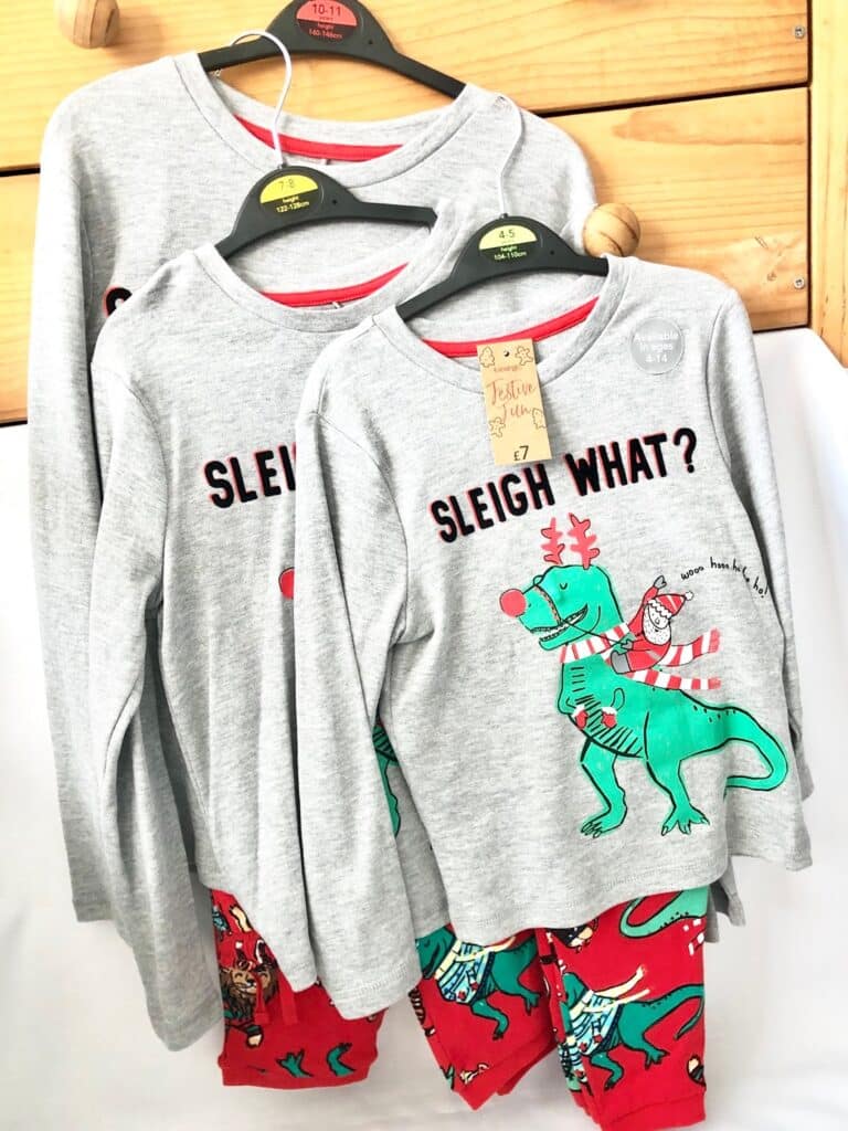 Three sets of children’s pyjamas are hanging, perfect for a 2018 Christmas or adding to a Christmas Eve Box. The grey tops feature a green dinosaur with festive details and “SLEIGH WHAT?” Matching red trousers display cheerful holiday prints.