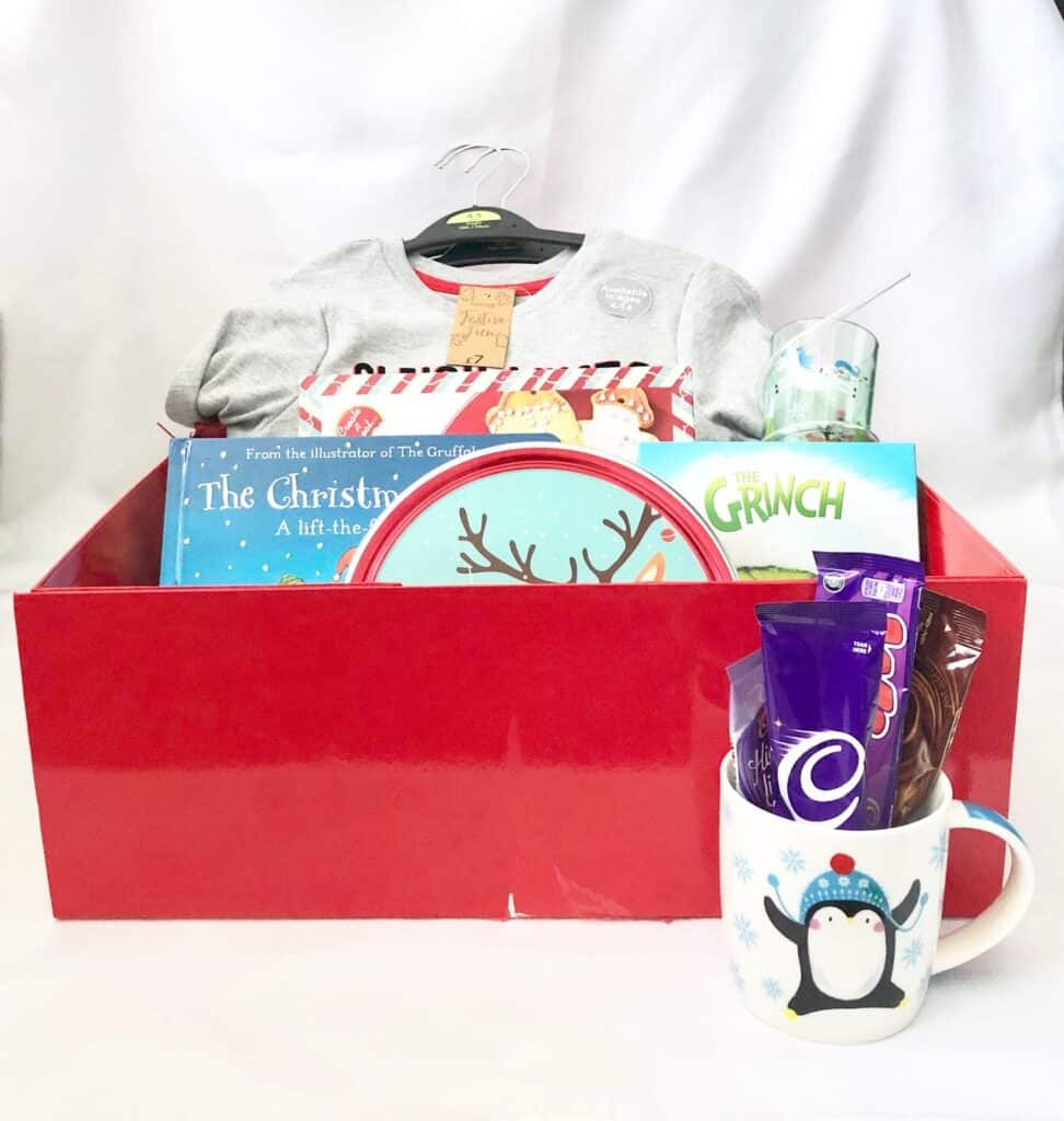 A festive Christmas Eve Box filled with holiday delights—a book, grey pyjamas, a decorative plate, a penguin mug holding snacks, and a drinking glass—all presented in a vibrant red box against a white background.