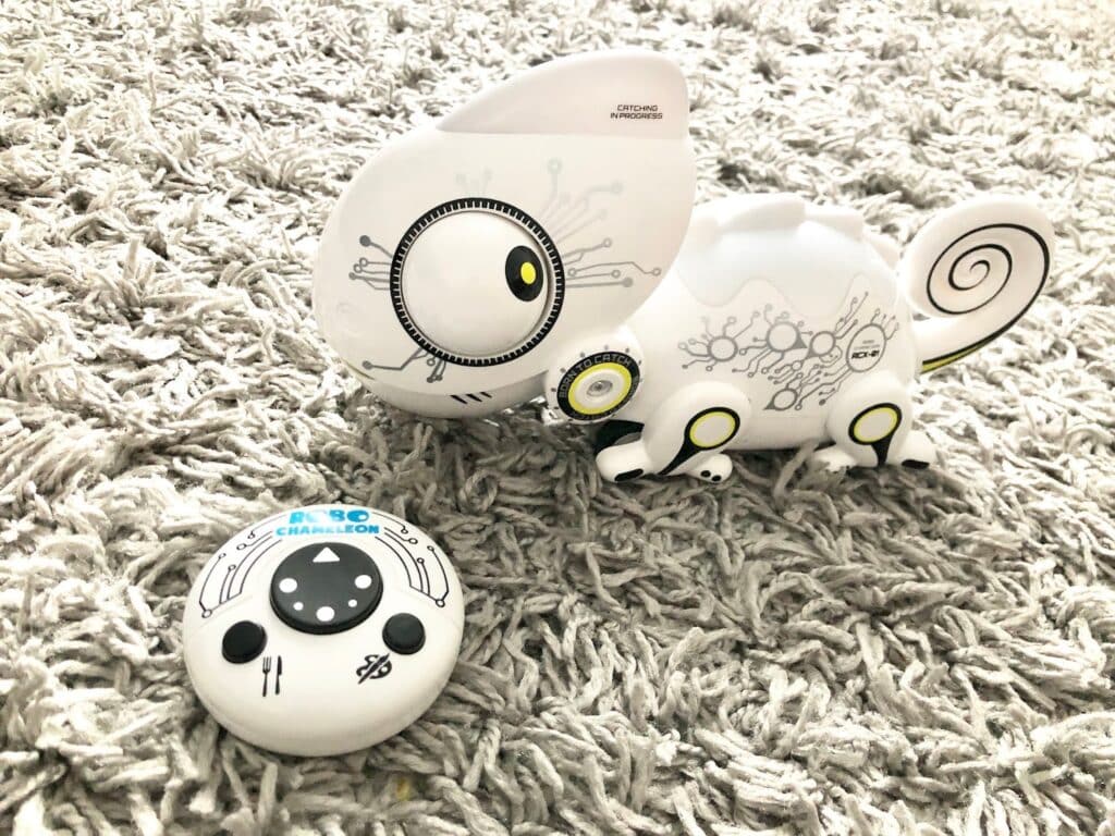 A white robotic chameleon toy with black circuit-like designs sits on a grey shag carpet next to a round remote control—perfect for Christmas fun and available from Wicked Uncle.
