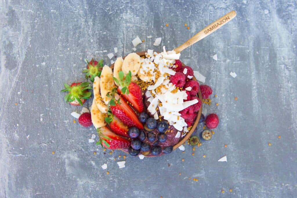 A colourful smoothie bowl, perfect as a breakfast recipe, is topped with bananas, strawberries, blueberries, raspberries, shredded coconut, and granola. A wooden spoon labelled "SAMBAZON" rests in the bowl on a grey textured background.