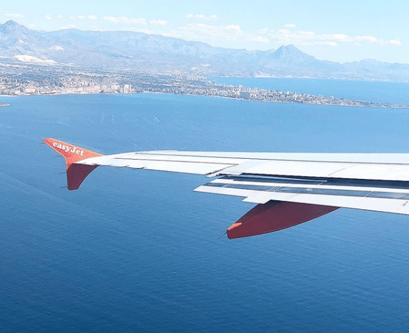 View from an aeroplane window showing the wing with “easyJet” branding, flying over a deep blue sea with a coastal city and mountains in the distance—perfect for anyone going abroad under a clear sky.