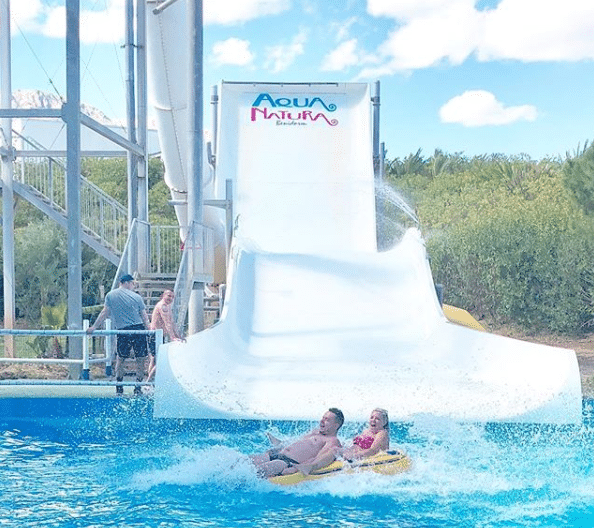 Two people on a yellow inflatable tube splash into a pool at the end of a large white water slide at Aqua Natura, enjoying a holiday adventure that feels like going abroad, with blue sky and greenery in the background.