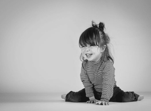 A young child with long hair wearing a striped shirt and dark trousers sits happily on the floor, smiling and looking slightly to the side in a black-and-white photo—a timeless family photography moment.