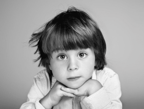 Black and white photo of a young child with straight hair resting their chin on their hands, looking directly at the camera with a calm, curious expression. A timeless piece of family photography, perfect as a Christmas present.