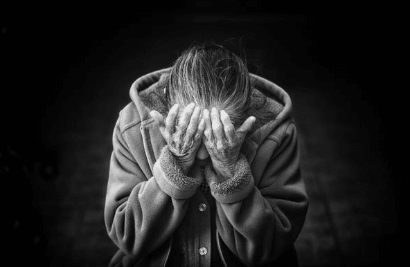 An elderly person in a coat sits with head bowed and hands covering their face, conveying distress. The black and white image with a dark background reflects the emotional challenges of ageing family member care, such as elderly incontinence.