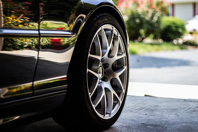Close-up of a shiny, silver alloy wheel and tyre on a dark-coloured car, parked on a driveway with greenery and a house in the blurred background—perfect for those who value car tyre care before travel.