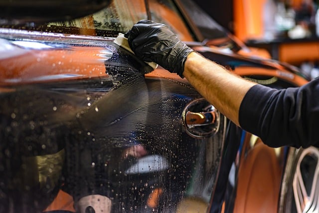 A person wearing a black glove cleans the side of a shiny black car with a white cloth, removing water and soap residue from the car door to help it look its best for an upcoming MOT test.