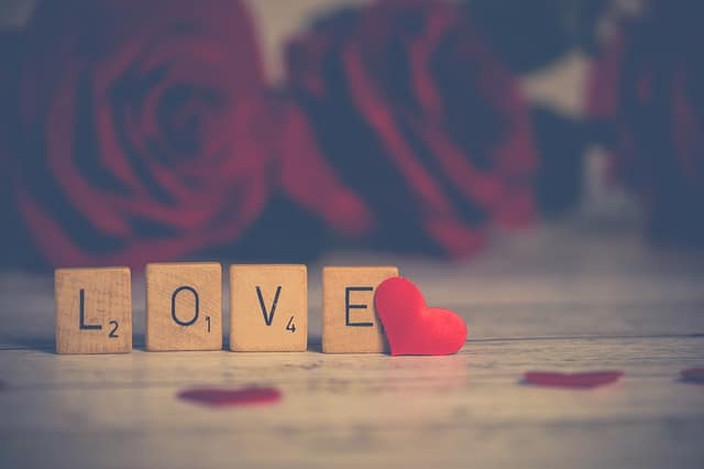 Scrabble tiles spelling "LOVE" are arranged on a wooden surface, with a small red heart and rose petals nearby—a charming display for anyone seeking alternative Valentine’s gestures to revive your love life. Blurred red roses appear in the background.