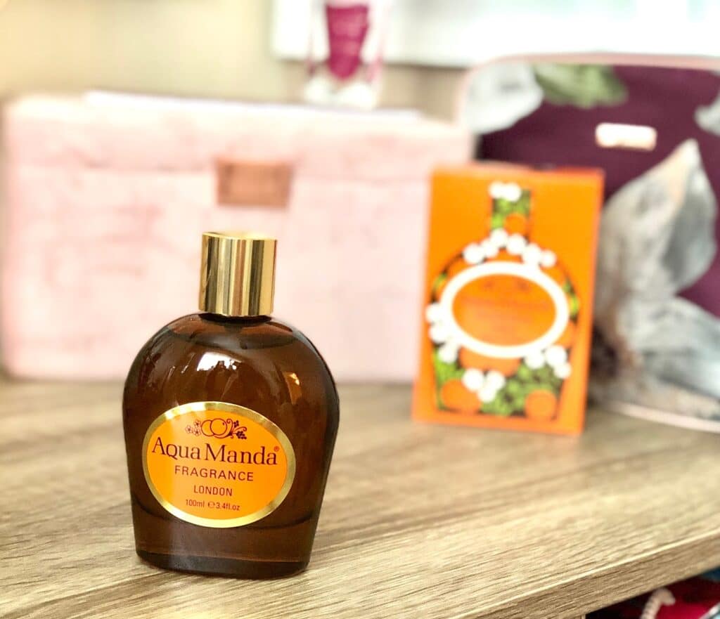 A brown glass bottle labelled "Aqua Manda Fragrance London" with a gold cap sits on a wooden surface, capturing the essence of vintage fragrances. In the background, an orange box and blurred pink and purple objects add to the retro charm.