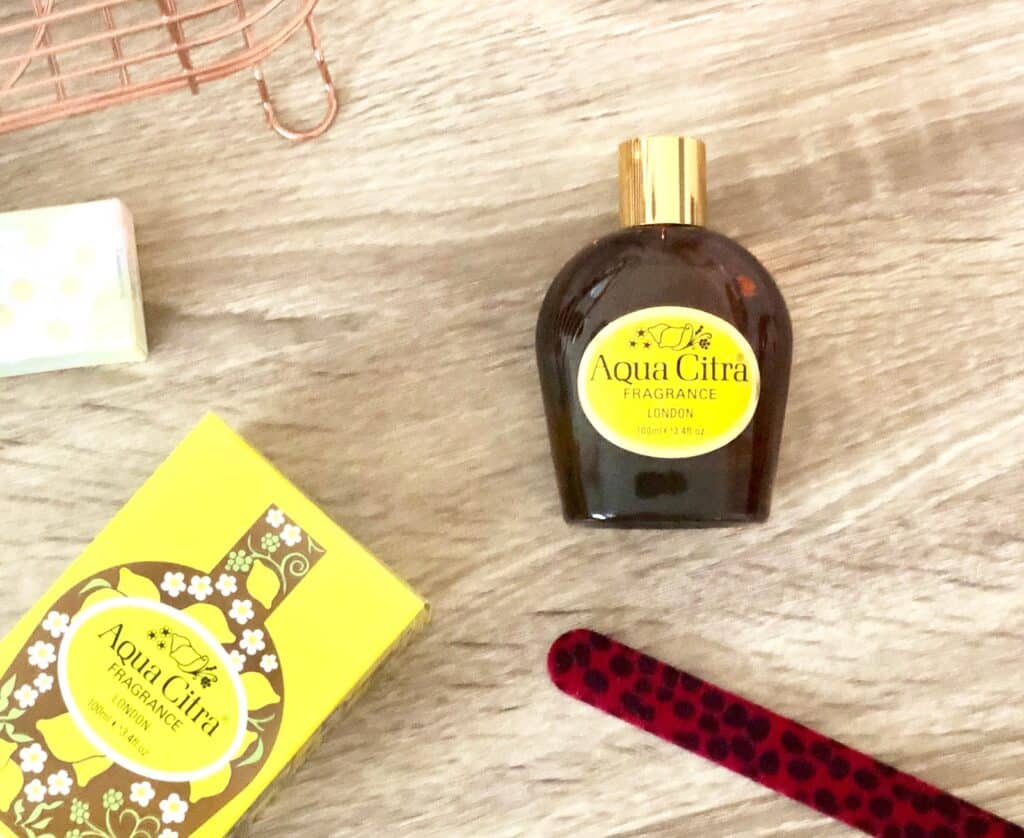 A bottle of Aqua Citra, one of the classic scents, with a gold cap sits on a wooden surface next to its yellow floral box. A pink nail file and part of a wire basket add charm to this nod to vintage fragrances.