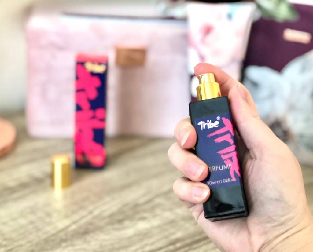 A hand holds a black perfume bottle labelled “Tribe” with pink design; this tribute to vintage fragrances sits beside a matching box, with blurred background objects on a light wooden surface.