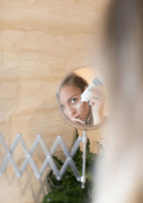 A woman’s face is reflected in a small round bathroom mirror as she applies skincare to her cheek, with a beige tiled wall and some green leaves in the background.